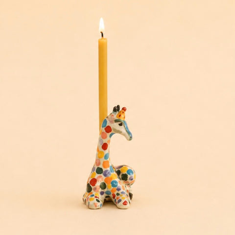 A lit yellow taper candle standing upright in a colorful giraffe-shaped holder against a warm beige background.