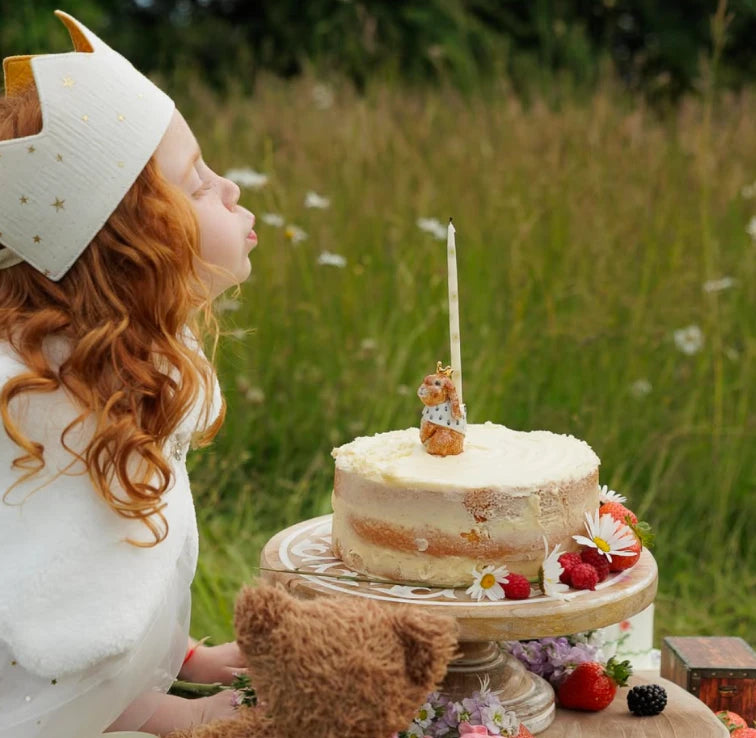 A child in a white outfit and matching hat blows a candle toward a frosted cake on a pedestal stand outdoors in a grassy field. A teddy bear sits at the base of the stand, with strawberries and flowers around the cake.