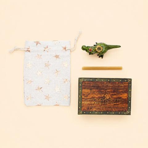 A flat-lay product shot of a small set on a warm cream background, including a light blue drawstring pouch with metallic star patterns, a green crocodile-shaped incense holder, a wooden incense stick, and a decorative wooden box labeled 'empfehl'.