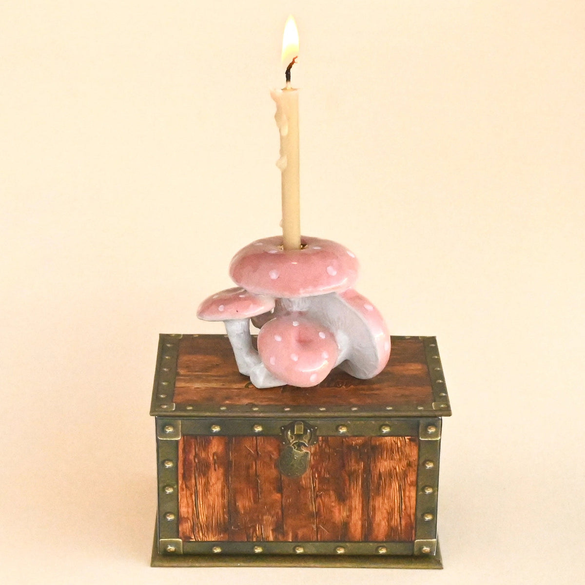 A decorative candle holder shaped like a small wooden chest with pink mushrooms and a lit candle inserted into the top, photographed against a plain beige background.