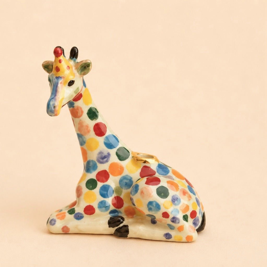 A ceramic giraffe figurine in a sitting position, decorated with colorful hand-painted polka dots in red, blue, green, yellow, and orange on an off-white base, set against a solid light beige background.