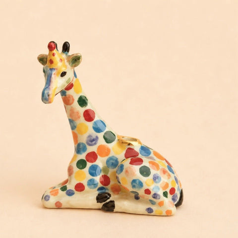 A ceramic giraffe figurine in a sitting position, decorated with colorful hand-painted polka dots in red, blue, green, yellow, and orange on an off-white base, set against a solid light beige background.