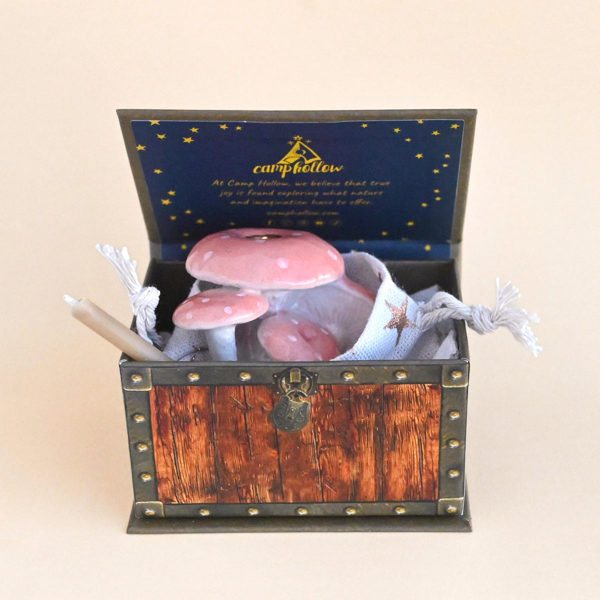 A miniature wooden treasure chest gift box with a dark blue starry lid printed with 'camphollow' and a candle inside, holding three pink-and-white polka-dot mushrooms and a white pouch, set against a solid beige background.