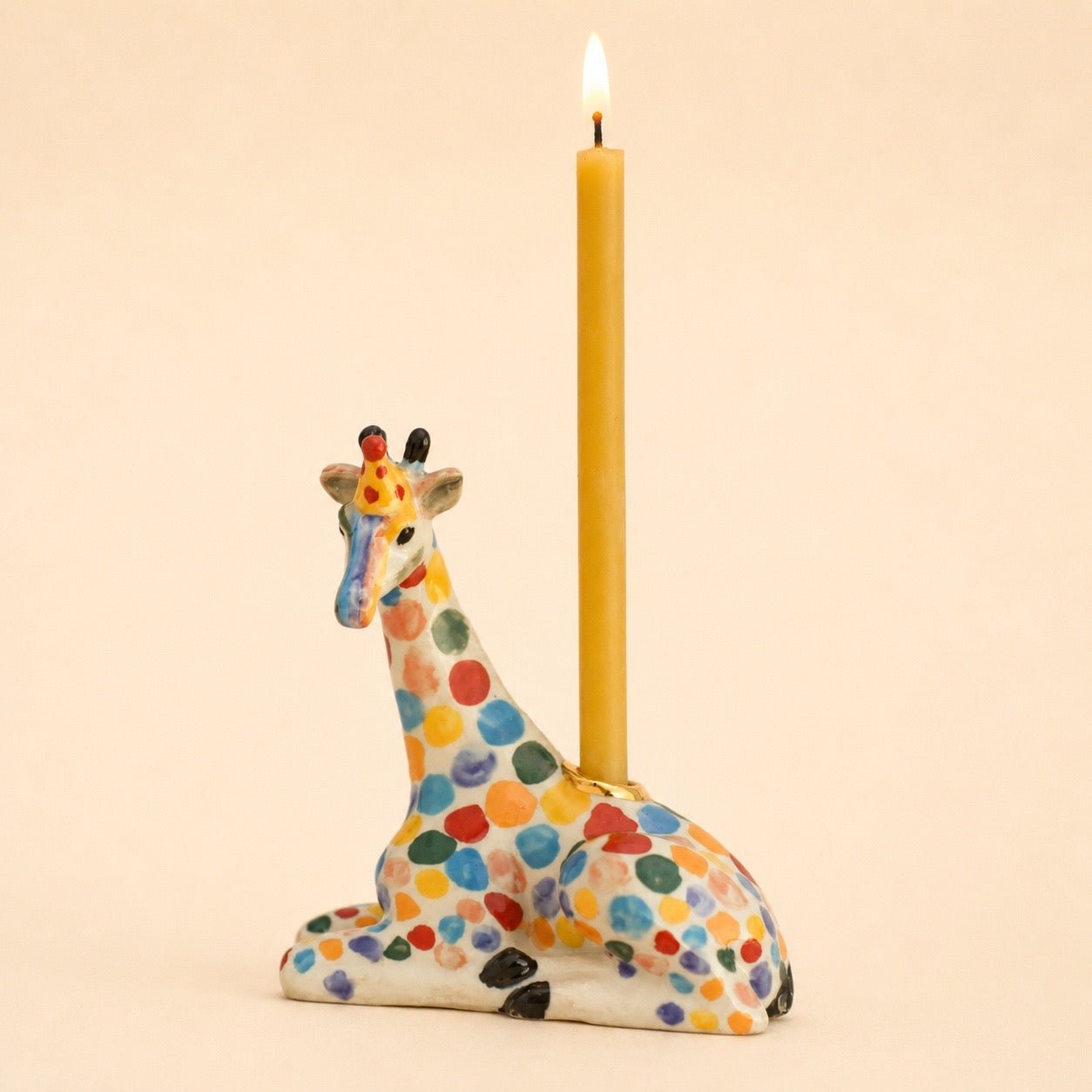 A ceramic giraffe-shaped candle holder with a colorful polka-dot pattern, holding a lit yellow taper candle against a solid beige background.