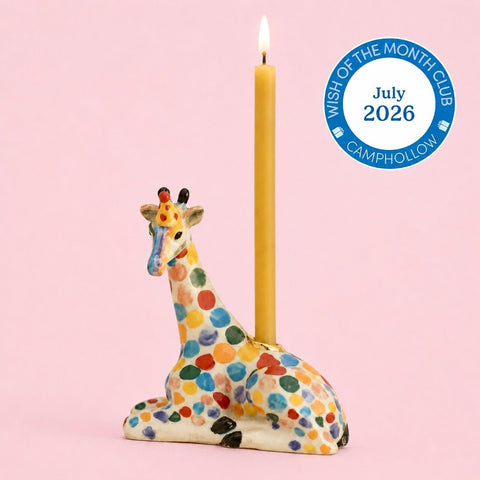 A ceramic giraffe candle holder with a lit yellow candle, decorated with colorful polka dots on a light pink background. A blue circular badge in the top right reads 'Wish of the Month Club July 2026 CAMPHOLLOW'.