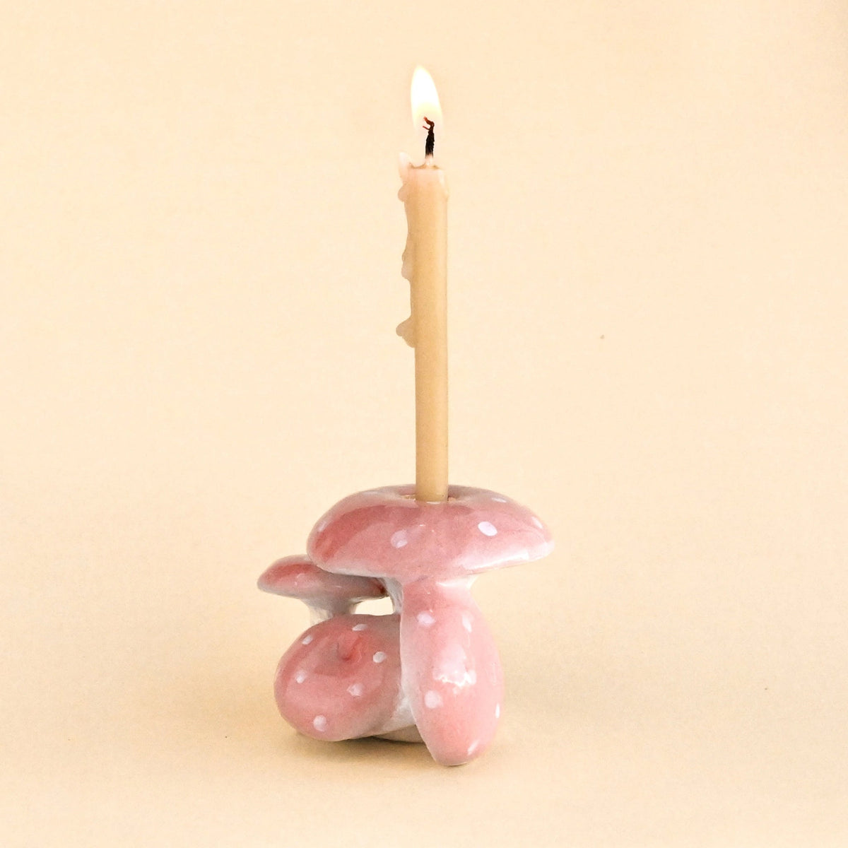 Front view of a pink mushroom-shaped candle holder with white polka dots, holding a lit beige candle, set against a solid beige background.