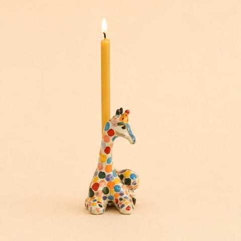 A lit yellow taper candle standing upright in a colorful giraffe-shaped holder against a warm beige background.