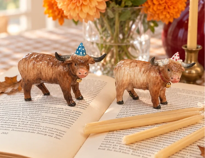 Two Highland cow birthday cake toppers on an open book beside autumn flowers and candles